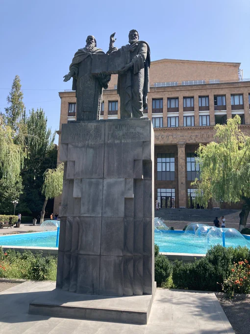 Statue of two scholars on a high cement plinth in front of a pool with fountains. Yerevan state university building in the background