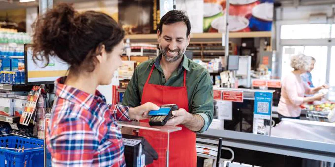 A younger woman holds her a payment card up to a cashier at a grocery store in the checkout line. The cashier is holding a card reader.