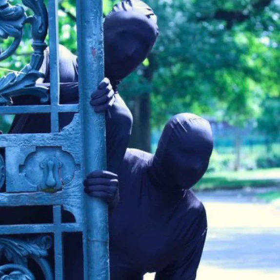 two actors peering from the side of a gate in a full body costume covering their face and hands, as pawns in a chess piece