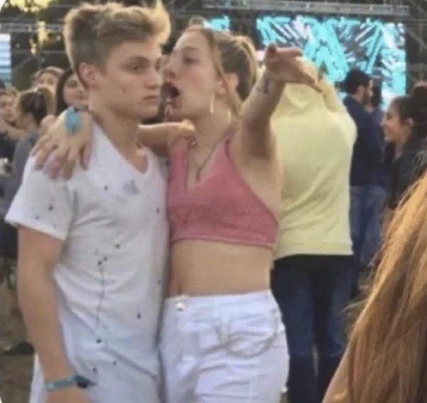 A person seeming to yell into the ear of another person, who has an uninterested expression on their face. The pair are in a crowd, with a stage in the background. It looks like they're at a concert. 