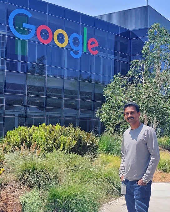 Johnson standing with hands in his pockets wearing a grey shirt and blue jeans in front of a blue Google office with Google written on the side. 
