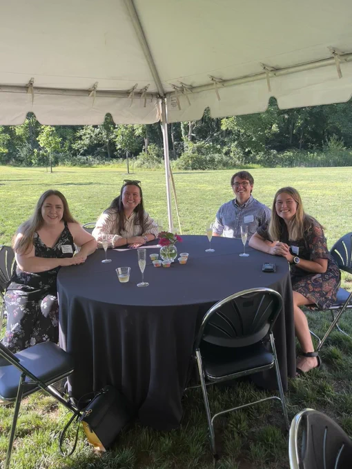 Baylee, Mallory, Noah, and Olivia sit around a table  under a tent outside