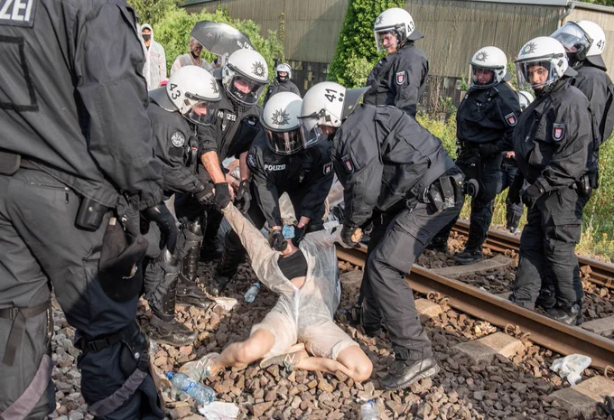 Vier Polizist*innen verdrehen den Kopf und die Hände eines/r Demonstrierenden von Ende Gelände auf Bahngleisen in Hamburg. Fünf Polizeikräfte stehen daneben, wovon eine Person zu lachen scheint. Foto: Jens Volle