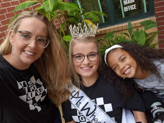 Sara in her crown and sash with her best friend and caregiver, Angelina, and Angelina’s daughter. They are wearing the Big Give Shirts and smiling at the camera.