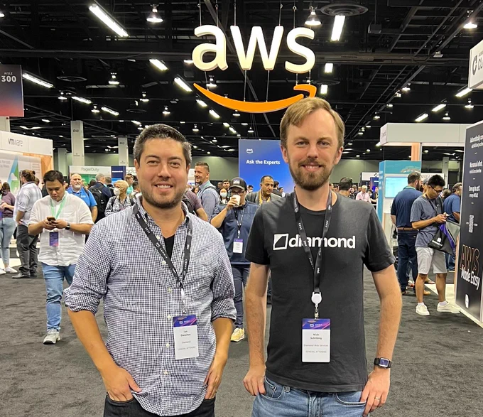 Two men standing under a sign that says "AWS." both men are wearing conference badges. the man on the right is wearing a shirt that says "diamond" on it.