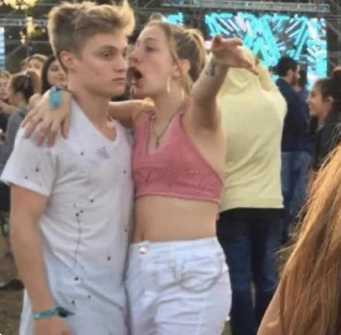 A young woman appears to be yelling into the ear of a young man. Her mouth is open. One arm is around him and one is up in the air. They appear to be at an outside festival.