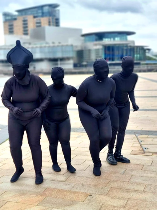 Four performers dressed in morph suits creep and sneak through media city piazza. One is wearing a chess pieces bishop headpiece