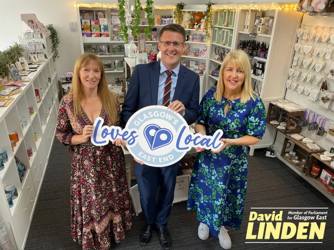 In the background of the picture is the interior of the gift shop and in the foreground David Linden MP is standing between two women from the Hoolit, all holding a large Glasgow East Loves Local sign.