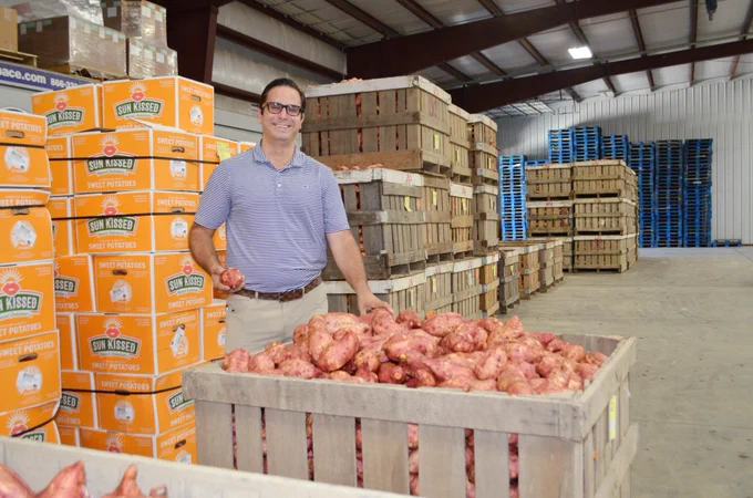 Photo of a business owner in front of boxes of sweet potatoes and behind a crate of sweet potatoes. 