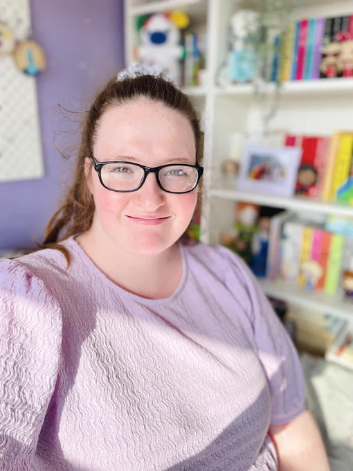A selfie of Shona, a young quite woman with long auburn hair. She is wearing a lilac too and glasses and smiling. A bookshelf can be seen in the background.