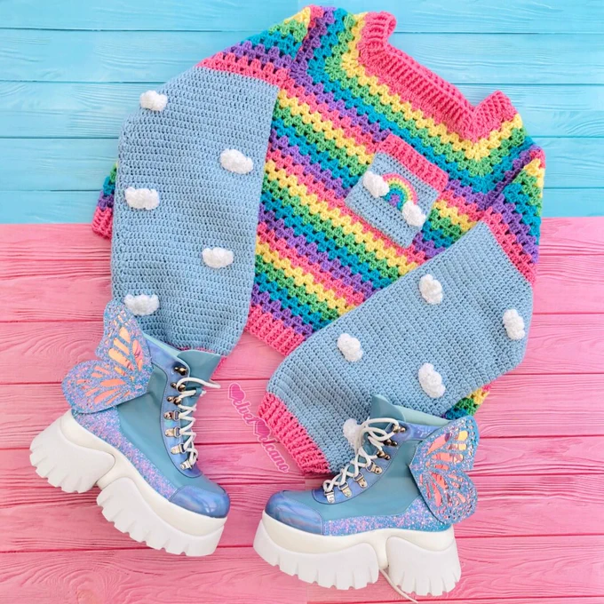 A crocheted jumper with pastel blue arms with white clouds on them and pink cuffs that also features a pastel rainbow striped chest/back section with a small blue square chest pocket with a rainbow appliqué on it and a pair of pastel blue chunky lace up boots with white soles and glittery holographic butterfly wings on the outer sides. The background is half turquoise tinted wood and half pink tinted wood