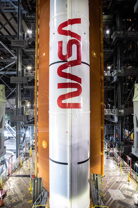 A view of a segment of the Space Launch System rocket’s orange core stage and its white solid rocket booster. The booster has the red NASA worm painted on it vertically.