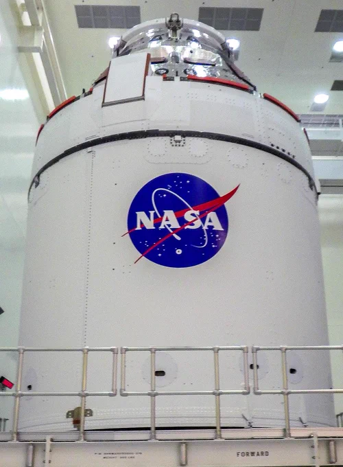 NASA’s Orion capsule sits atop its European-built servicing module. Orion has silver, reflective paneling and the module is white. Together they are upright in a processing facility. The NASA emblem, blue with a red streak over and behind the letters N A S A, is painted on this side of the servicing module. 