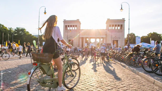 Photograph of a woman on a green bicycle cycling off into the sunset, which is visible behind a large gate in Munich. Many cyclists are seen in the photo. 
