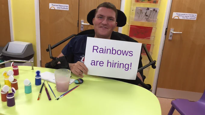 A young man sat in a wheelchair smiling, saw he holds a sign which reads "Rainbows are hiring!", there's paint pots and paint brushes on the table in front of him.