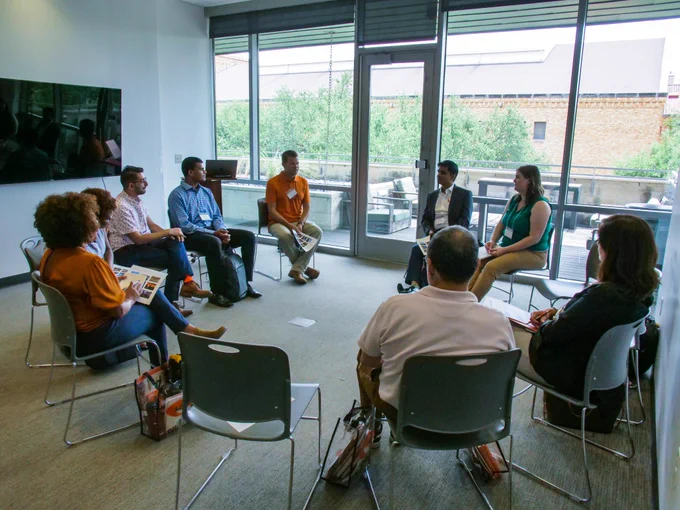 Group of NFS 22 participants sitting in a circle of chairs and having a discussion. Glass windows are in the back of the photo with trees visible and a building. 