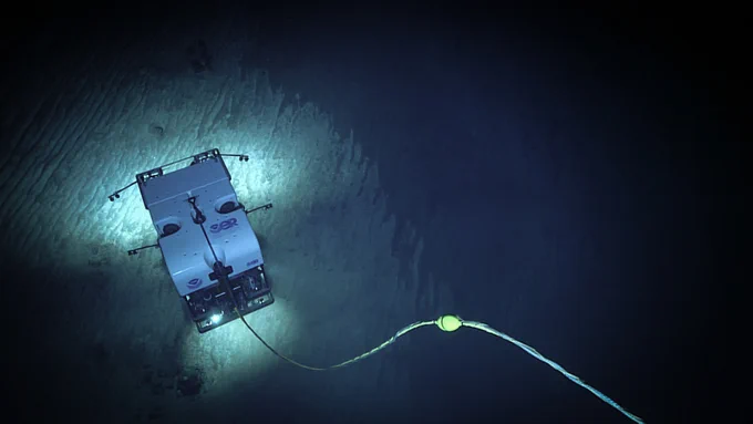 A remotely operated vehicle -- a boxy underwater robot about the size of a van -- is seen from above as it explores the seafloor, which is illuminated by its lights.