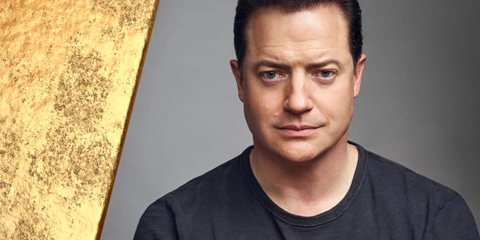 A close-up of Brendan Fraser wearing a dark grey shirt beside a gold bar. He stares directly into the camera with one eyebrow slightly raised and his lips pressed together.