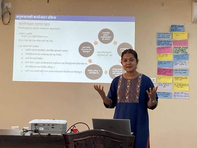Lady in a blue sari stands in from a wall with a diagram and Nepali writing projected onto it