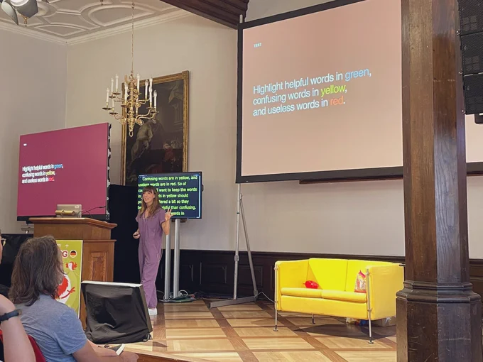 Sophie Tahran on stage; a slide in the background shows „Highlight helpful words in green, confusing words in yellow, and useless words in red.“