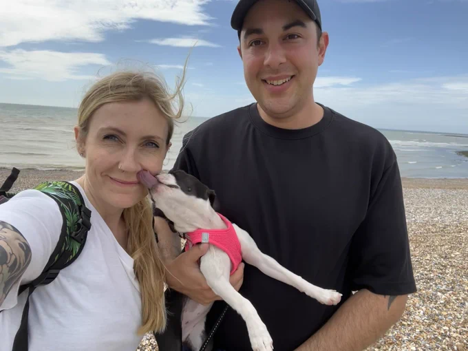 Jon and Kelly selfie. Jon is holding Boston terrier puppy, Freya, who is giving Kelly a kiss. The background is the sea and pebbly shore
