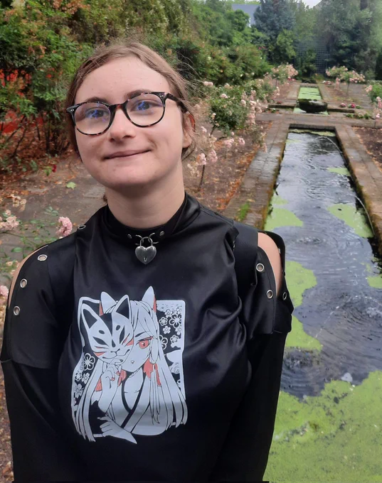 Jeune femme portant des lunettes et un haut noir, souriante. Paysage vegetal en arriere plan, avec des bassins rectagulaires.