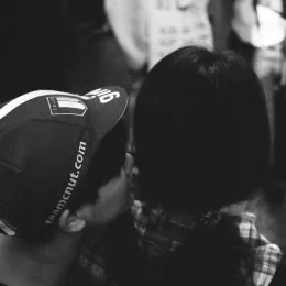 A black and white image of the back of two people's heads. The person on the left is wearing a cycling cap and whispering in the ear of the person on the right. 