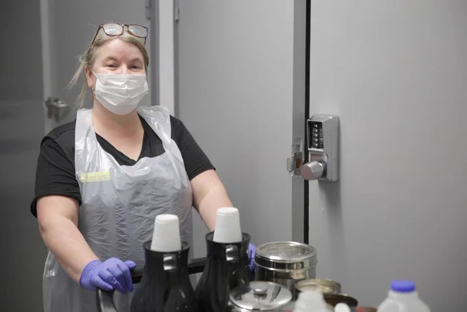 A member of staff wearing an apron, mask, and gloves while delivering drinks to patients.