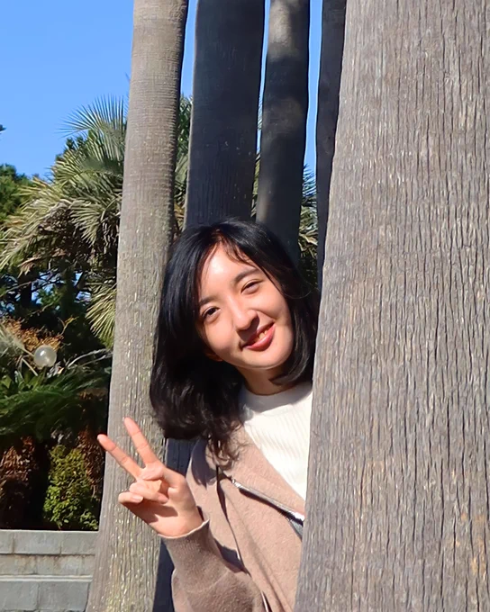 Sumin wearing a tan jacket and white shirt leaning out from 
behind a tree smiling holding up a peace sign.