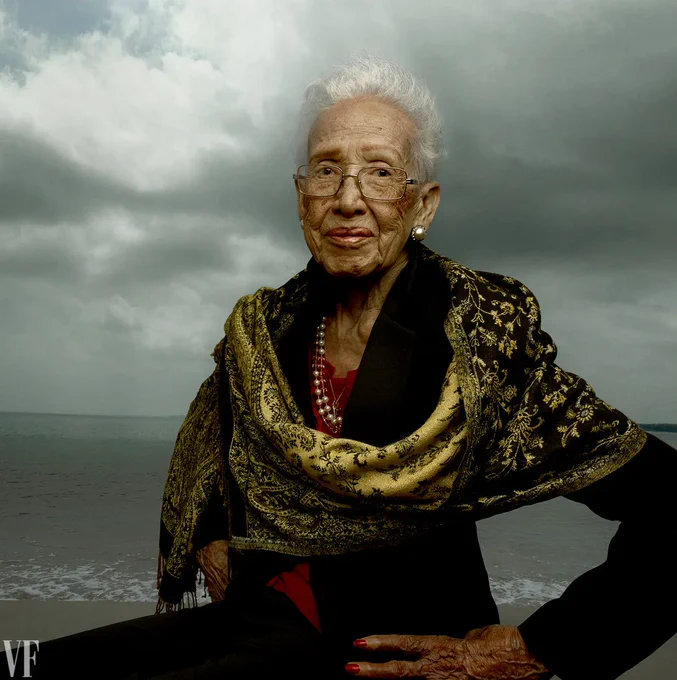 Portrait of African American woman mathematician, Katherine Johnson. Johnson is in the foreground in a black pant suit and claret blouse. She has a manicured hand with red nail polish placed on her right hip. The has a black and gold shawl draped across her shoulders and two strand pearls around her neck. She is wearing pearl earrings. She stands on the beach in front of the ocean in Hampton Virginia, at dusk. 
credit: Annie Liebowitz, Vanity Fair (2016)