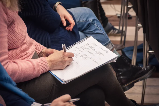 People are sat together and one is writing on a notepad, all gathered in a creative environment.