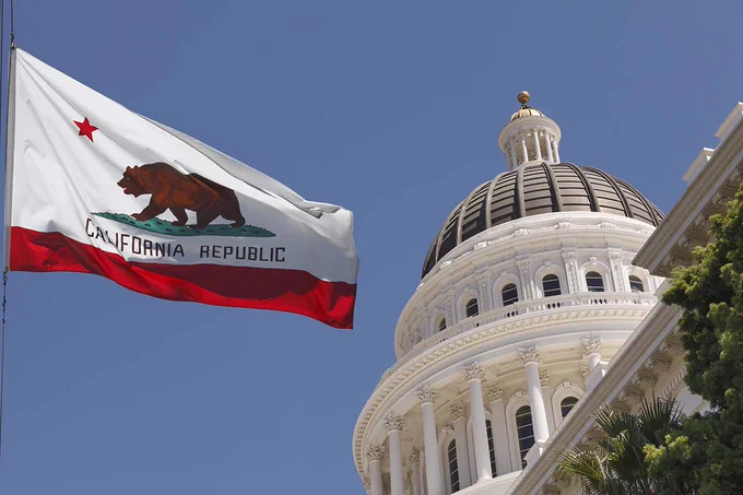 The California flag is flying next to the capitol rotunda in Sacramento, with clear blue sky in the background.