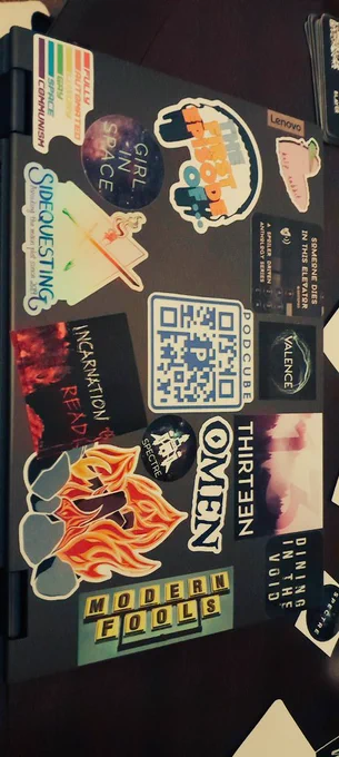 Pictured: top of a laptop computer, absolutely coated in stickers. The stickers are mostly of various audio fiction shows. Listed from left to right, top to bottom they are: Kelp Rabbit (artist), Someone Dies in this Elevator, Valence, Thirteen, Dining in the Void, The First Episode Of, PodCube, Omen, Spectre, Modern Fools (band), Fully Automated Luxury Gay Space Communism (art), Girl in Space, Sidequesting, Incarnation Read, Fire (art). 