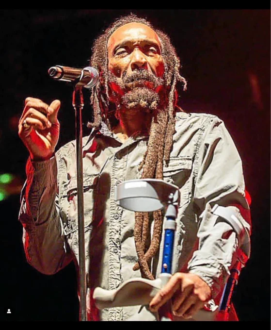 Homem negro com dreadlocks longos em frente a um microfone e a uma muleta 