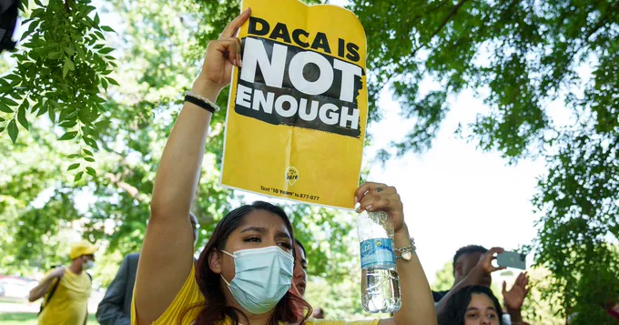 A protestor wearing a mask and holding a water bottle and sign overhead that reads "DACA is not enough." There are few other people in the background and lots of foliage.