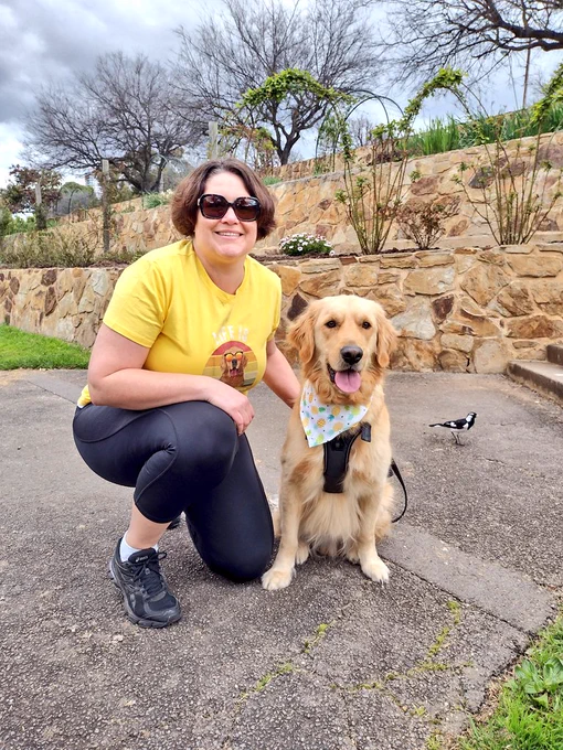 A white woman with medium length brown hair and sunglasses is crouching in front of a rock wall. She's wearing a yellow tshirt with an image of a golden retriever wearing sunglasses. Her golden retriever Panko is sitting next to her. He's wearing a bandana with pineapples on it and his tongue is hanging out