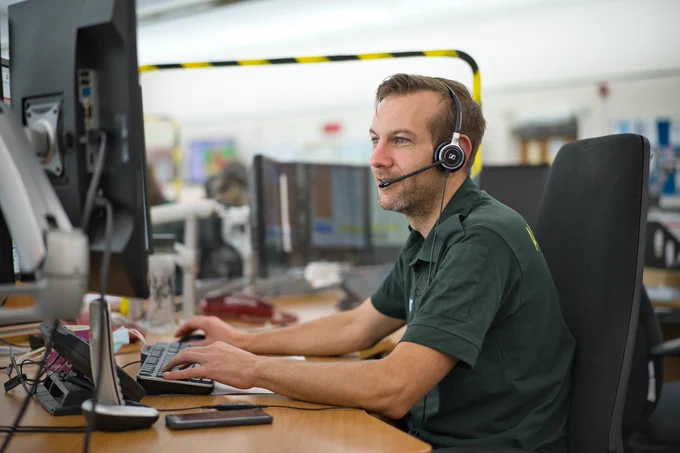 Male call handler in green uniform talking into a headset whilst typing on a computer keyboard. 