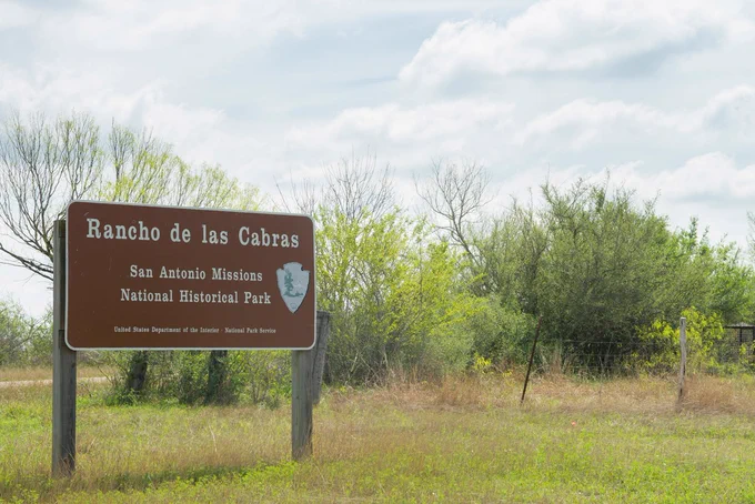 Wild Texas ranchland on a cloudy day with a brown sign in front reading “Rancho de las Cabras, San Antonio Missions National Historic Park, United States Department of the Interior, National Park Service”