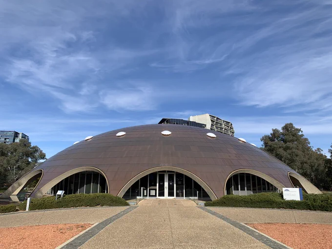 The Shine Dome is shown with archways extending around the base of a circular building covered in copper tiles, under a blue sky with light clouds.  