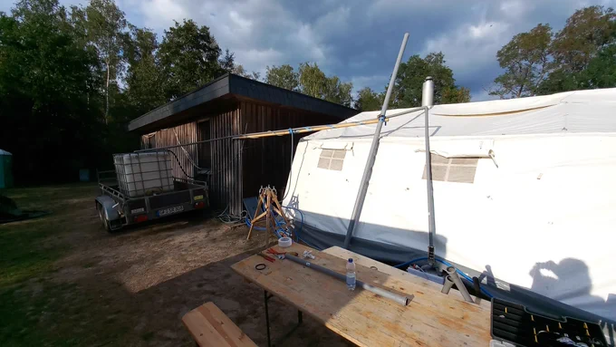 Abflussrohr ist an einem Rundholz befestigt, welches am Haus und an einem dicken, ans Zelt gelehnten Abflussrohr aufgehängt ist. Das Rohr mündet nun in einen IBC auf einem Anhänger.