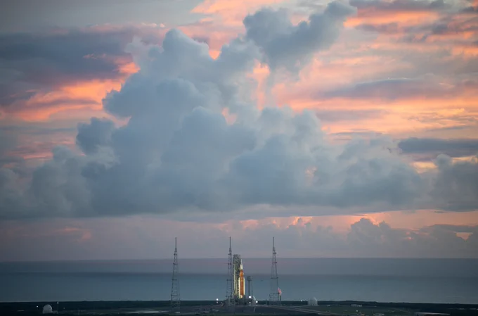A panoramic view of the Space Launch System rocket at the launch pad a few miles away, getting illuminated during sunrise. Its 32-story orange core stage is flanked by two white rocket boosters. In the background is the Atlantic Ocean and towering grayish-purple clouds dominate the image. The sky is aglow with orange hues between the clouds.
