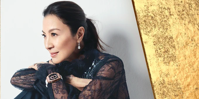Michelle Yeoh sits with her arms folded across her knees, gazing off to the left in front of a white background. She wears a gold watch, silver earrings and a black top with sheer, lacy sleeves. She has a small smile on her face, and her hair is pulled back in a ponytail.