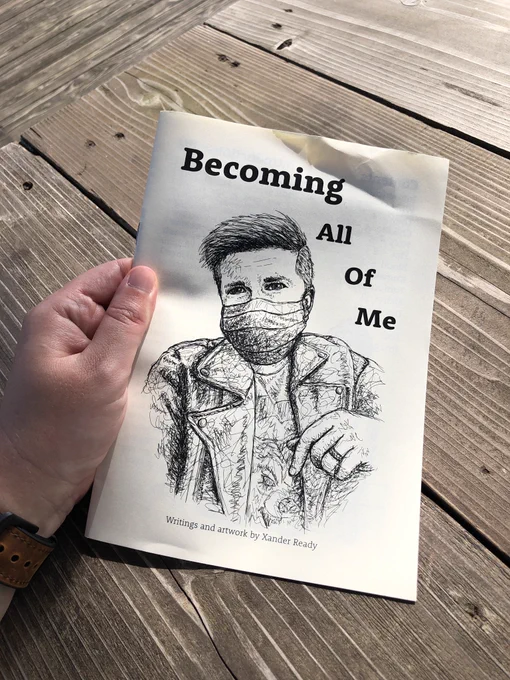 A photo of a hand holding an off-white paper booklet titled “Becoming All of Me” just above a rough wooden table, subtitled “Writings and artwork by Xander Ready.” Below the title is a detailed digital ink bust sketch of a young white man with short hair wearing an accordion-style cloth mask and a leather jacket, looking at the camera with a confident expression.