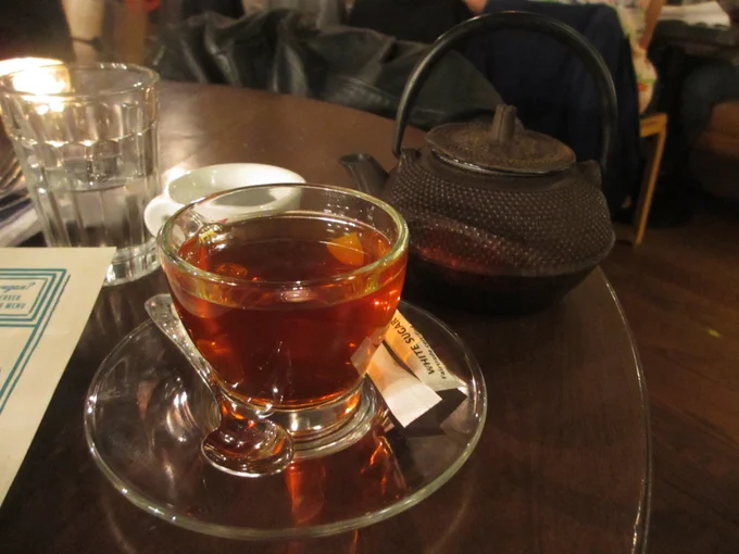 A see-through cup of black tea sits on a table, with an iron tea kettle in the background. 