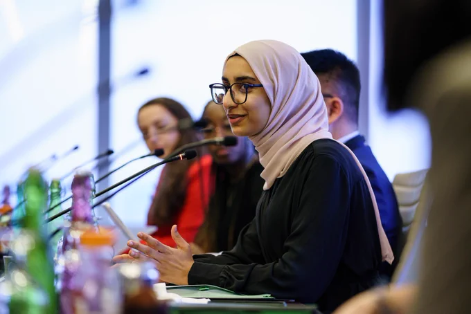 A young person, who is a member of the Prime Minister Youth Council, is sitting and speaking into a microphone. Others are sitting beside them.