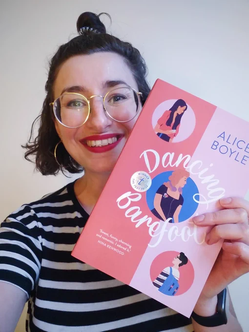 Alice Boyle, a light-skinned woman with dark hair, glasses, and wearing a striped t-shirt, is holding a copy of Dancing Barefoot and looking excited. The book has a reddish-pink and lilac cover and illustrations of the three main characters on the front. There's also a metallic silver stamp that shows that the book won the Text Prize.