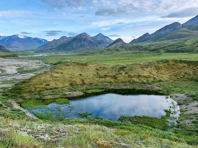 A small pond sits within a large open tundra area. In the distance are tall and steep mountains.