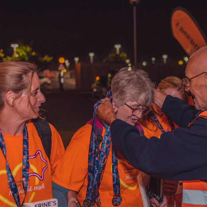 Two midnight walkers wearing orange t-shirts having medals placed over their heads.