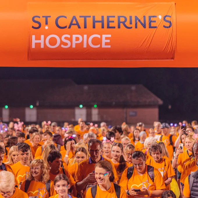Large group of people all wearing orange t-shirts, standing under a 'St Catherine's Hospice' inflatable start line. 