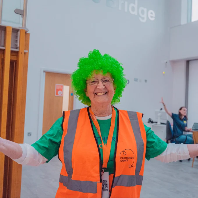 Volunteer in an orange high vis jacket and green wig, smiling to the camera. 